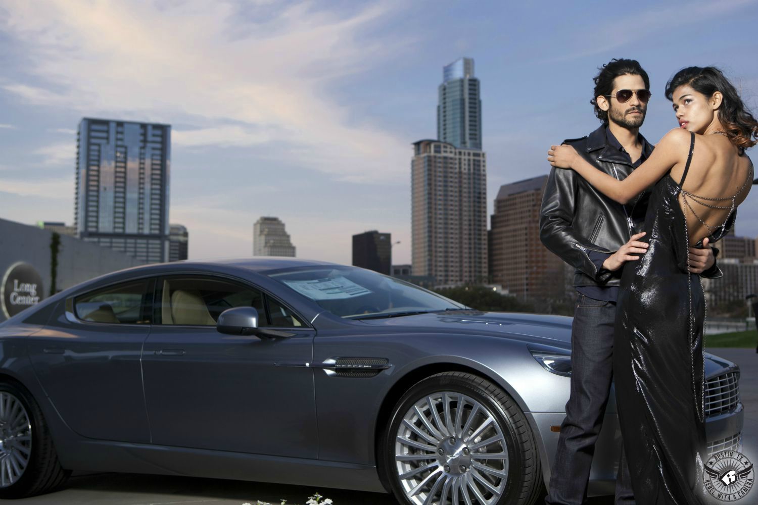 male and female fashion model embracing wearing leather coat and vinyl dress next to an aston martin with the city of austin in the background where you can see austin 360